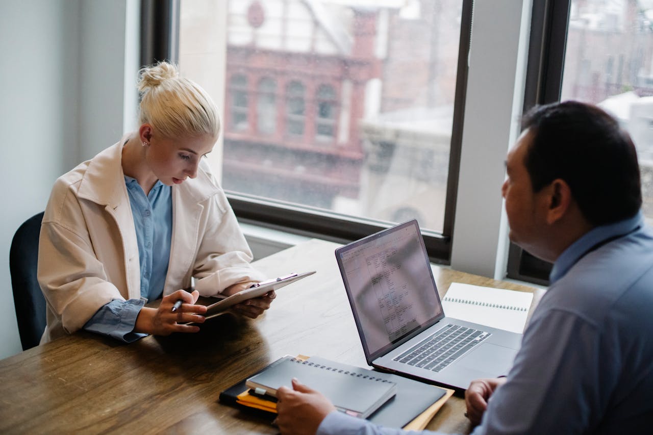 footer-02 From above of young female worker wearing modern coat analyzing details of project with colleague while sitting together at wooden desk with laptop and documents