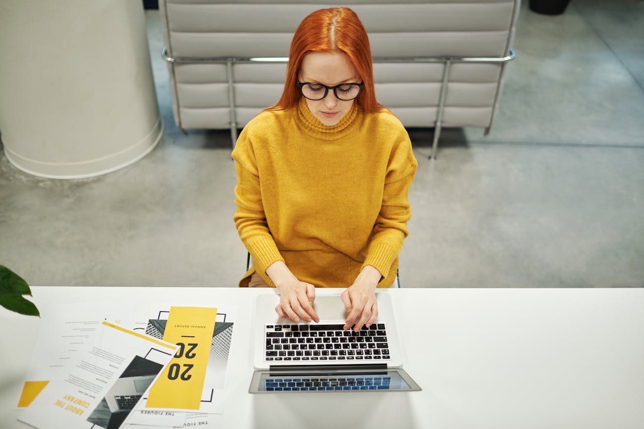 services-04 Redhead woman in a yellow sweater typing on a laptop in a modern office setting.