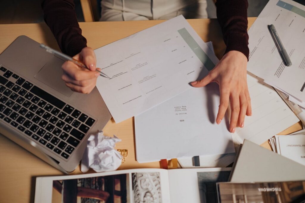 A person taking notes and working on a laptop surrounded by documents on a desk.